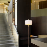 Danona P Fabric Drum Floor Lamp with a white pleated shade on a slim metal stem, shown beside a staircase in a modern dark-panelled interior.