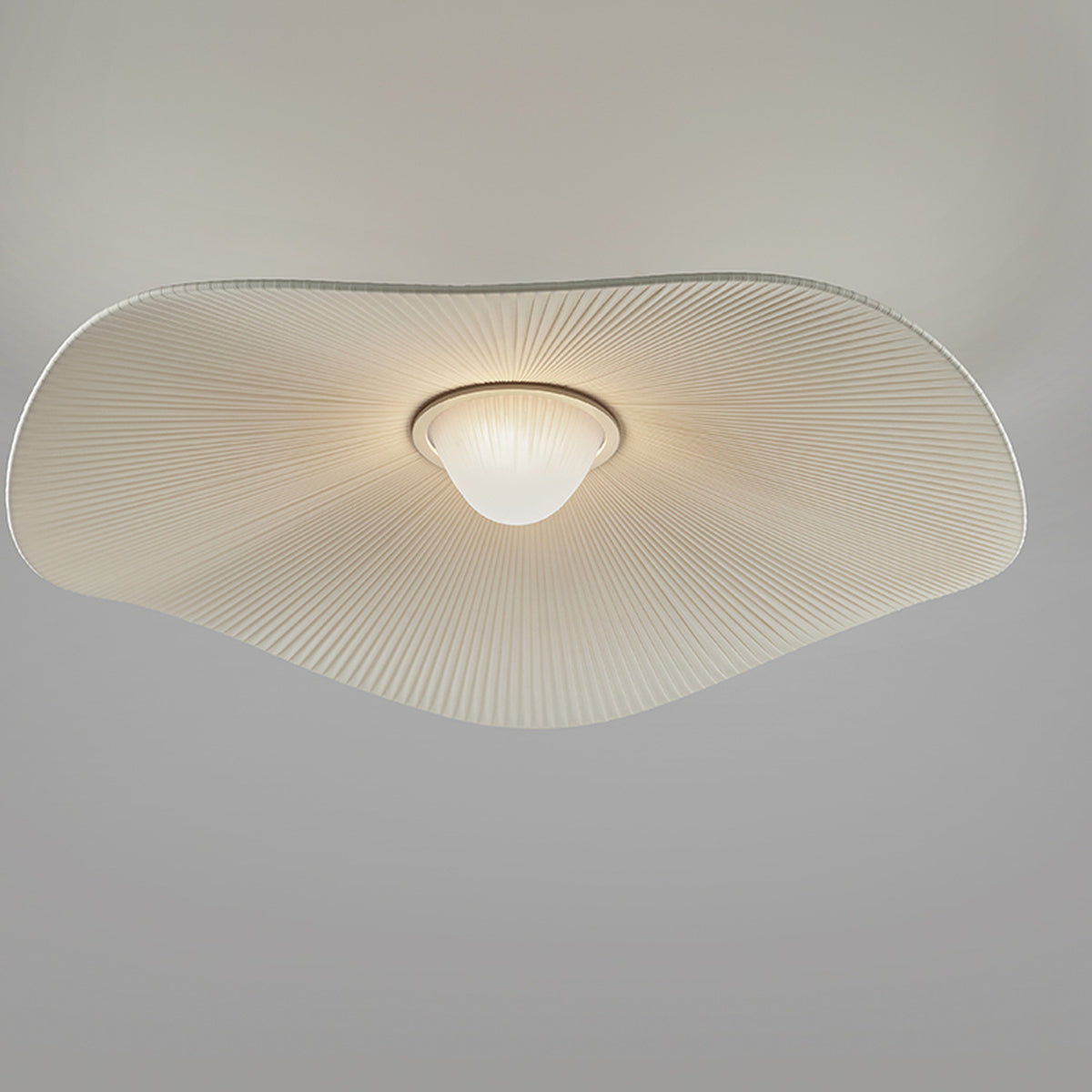 Mediterrània PF/105 ceiling light with a wide, wavy white translucent pleated ribbon shade and a central frosted glass diffuser, shown glowing against a plain grey background.