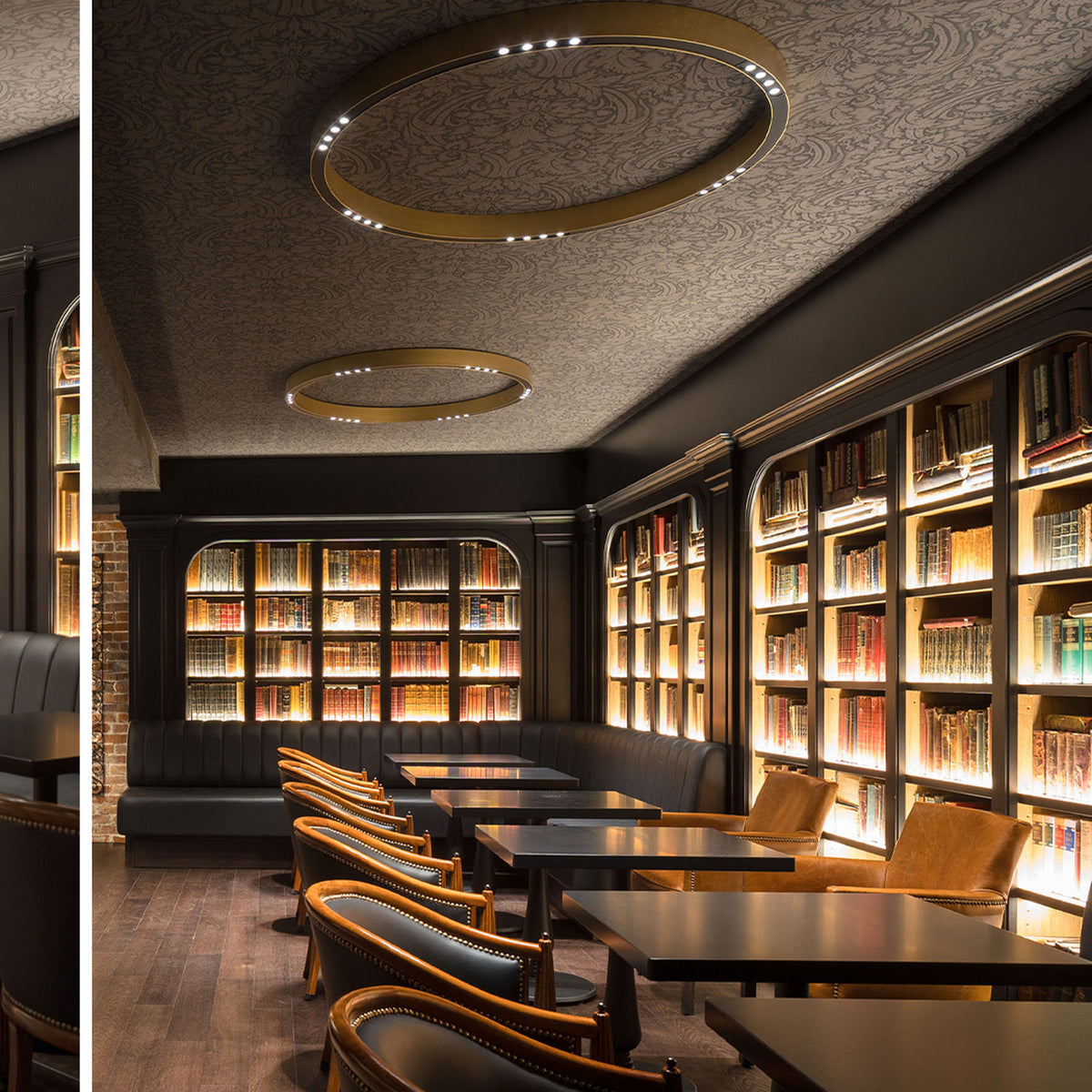 R2 C120 Aluminium Ring LED Ceiling Light in brass finish, two large circular fixtures with integrated LED points, shown above a bookshelf-lined lounge with dark wood tables.