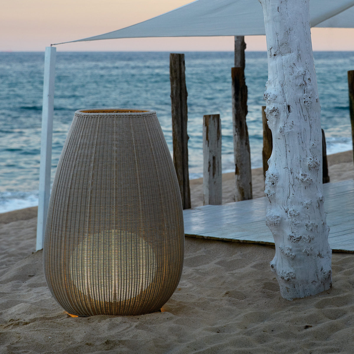 Amphora 03 Woven LED Outdoor Floor Lamp in beige, tall tapered woven body on short legs, glowing warmly on a sandy beach setting near the sea.