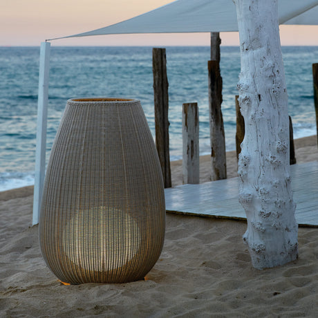 Amphora 03 Woven LED Outdoor Floor Lamp in beige, tall tapered woven body on short legs, glowing warmly on a sandy beach setting near the sea.