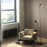Speers F 2 Light LED Floor Lamp in matte black with two dome-shaped heads on slim stems and a round base, shown beside a beige armchair in a living room.