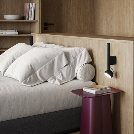 Berry W Aluminium LED wall light in matte black with a slim upright stem and round adjustable dome head, mounted beside a bed in a wood-panelled bedroom.