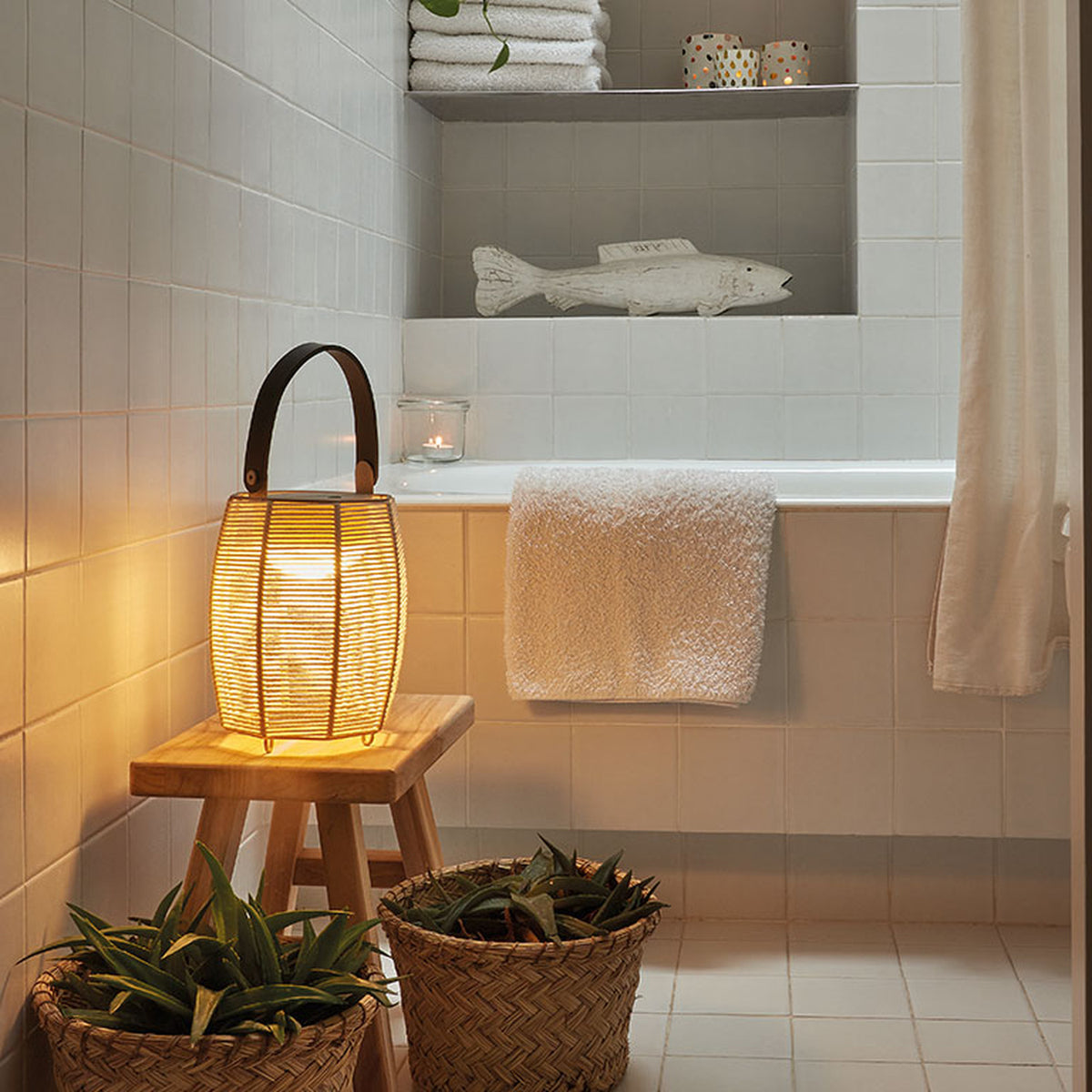 Tanit M/29/R rechargeable table lamp with brown woven fabric shade and curved handle, lit on a wooden stool in a bathroom beside towels and plants.