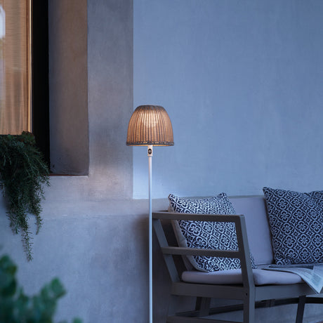 Atticus P/114/R rechargeable outdoor floor lamp with woven beige dome shade on a slim white pole, shown lit beside an outdoor sofa with patterned cushions.
