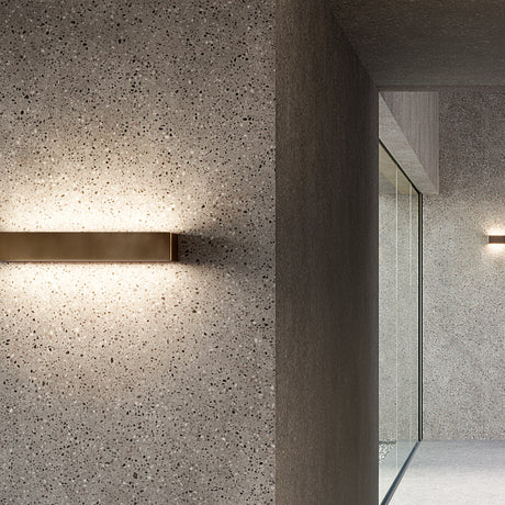 Toy S LED Wall Light 43.7cm in bronze, a slim rectangular bar fixture mounted on a textured terrazzo wall, casting warm uplight and downlight along a hallway.