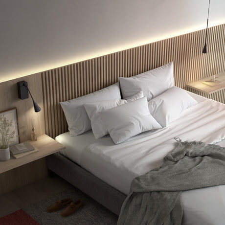 Skybell Plus A/01 wall light in Ebony Black, mounted above a bedside shelf with a slim iron rod arm, small bell-shaped head and integrated LED, shown beside a slatted wood bedroom wall.