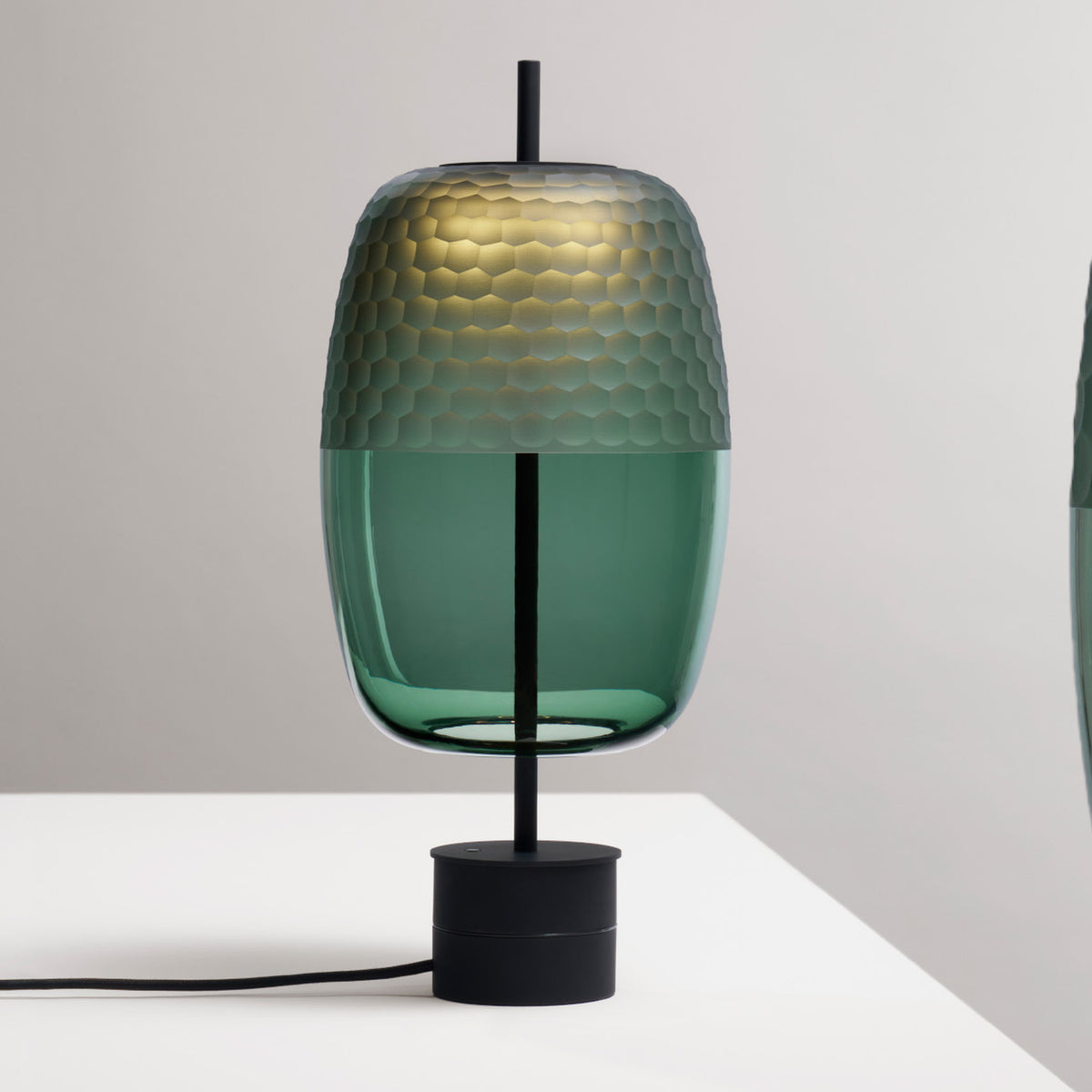 Adamas 23 table lamp with a lantern-shaped forest green glass shade, honeycomb-faceted top and clear base, on a slim black rod with round base, set on a light background.