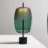 Adamas 23 table lamp with a lantern-shaped forest green glass shade, honeycomb-faceted top and clear base, on a slim black rod with round base, set on a light background.