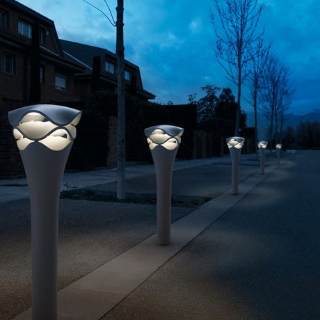 Cornet B/77 Tiered LED Bollard Light in stone grey lining a path at dusk, slim flared stem topped with a wavy layered head glowing warm white.