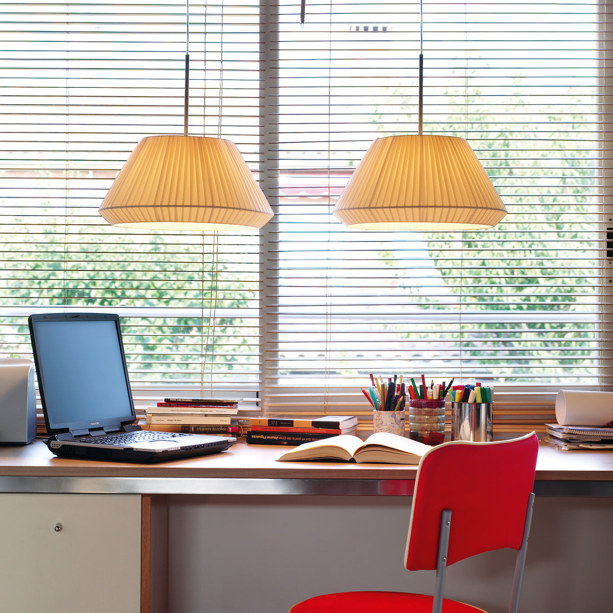 Two Mei 38 Ribbon Cone pendant lights with cream pleated ribbon cone shades, hung above a home desk with a laptop, books and a red chair.