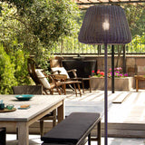 Ralph 50 outdoor floor lamp with woven metallic brown cone shade on a slim stem and tripod base, shown illuminated on a patio beside outdoor dining furniture.