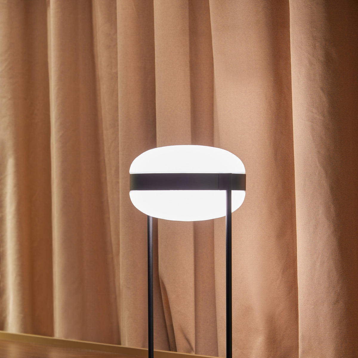 Beaded T Glass LED Table Lamp with an opal white, flattened spherical shade held by a dark central band between two slim black rods, set against a tan curtain backdrop.