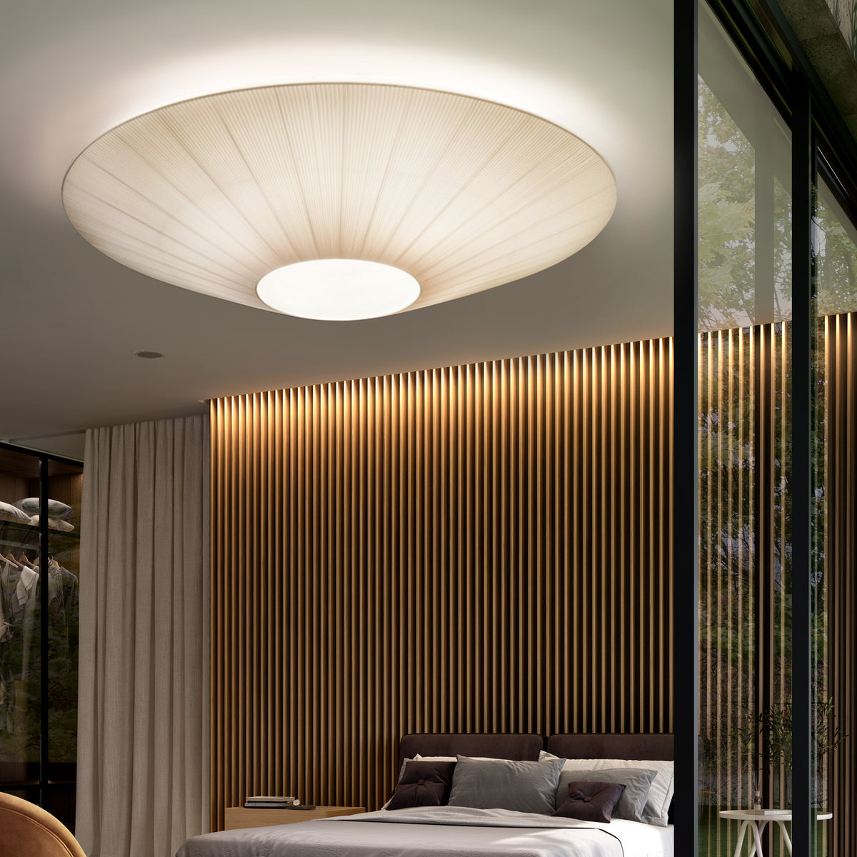 Siam 150 Ribbon ceiling light with a wide, pleated cream translucent shade and round white centre diffuser, shown illuminated above a bed in a modern bedroom with wood-slat panelling.