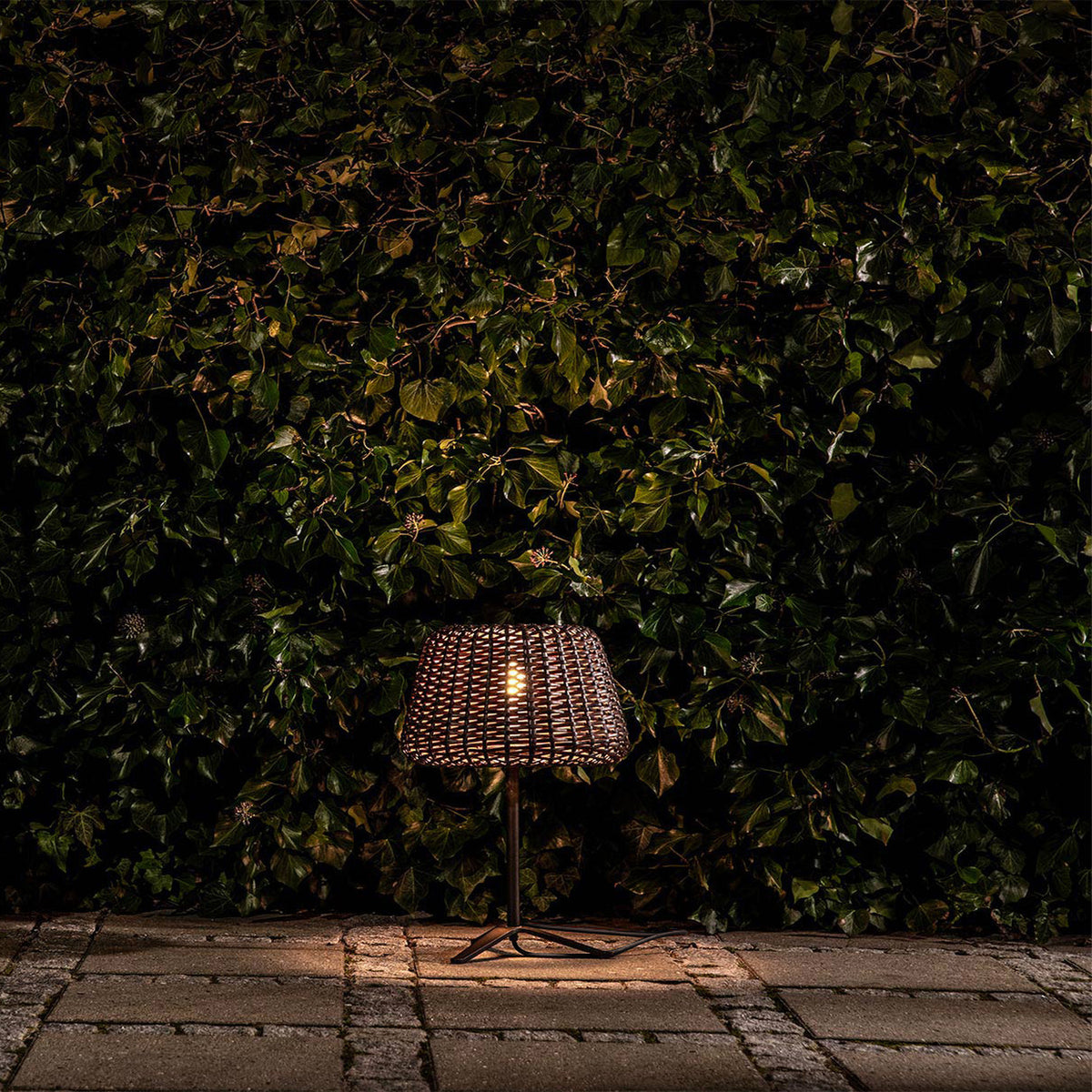 Ralph Woven Cone Outdoor Table Lamp in Metallic Brown, with tapered rattan-effect shade, slim stem and cross-shaped base, glowing warmly on a paved patio against a dark green hedge.