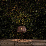 Ralph Woven Cone Outdoor Table Lamp in Metallic Brown, with tapered rattan-effect shade, slim stem and cross-shaped base, glowing warmly on a paved patio against a dark green hedge.