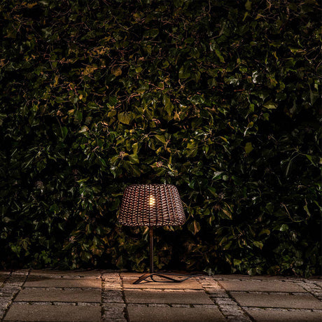 Ralph Woven Cone Outdoor Table Lamp in Metallic Brown, with tapered rattan-effect shade, slim stem and cross-shaped base, glowing warmly on a paved patio against a dark green hedge.