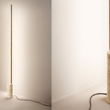 Hilow Line Travertine LED floor lamp with a slim brass-toned aluminium rod rising from a round travertine base, shown against a plain light wall.