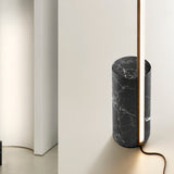 Hilow Line floor lamp with a slim Brushed Champagne aluminium rod rising from a cylindrical Nero Marquina marble base, shown against a neutral wall interior.