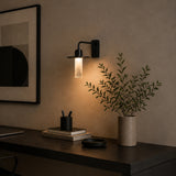 Beira W Glass LED Wall Light in matte black with a slim rectangular plate, downward arm, flat disc and frosted diffuser tube, shown glowing warm white above a desk with books and a plant.