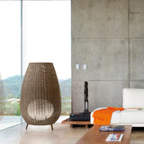 Amphora 03 Rattan LED Outdoor Floor Lamp in brown woven synthetic fibre, tall amphora-shaped shade on three short legs, shown beside a sofa in a living room with concrete wall.