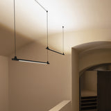 BIMU S150 Dep linear LED pendant lights in matte black, slim tube fixtures suspended on fine cables at an angle in a vaulted, warm-toned interior.
