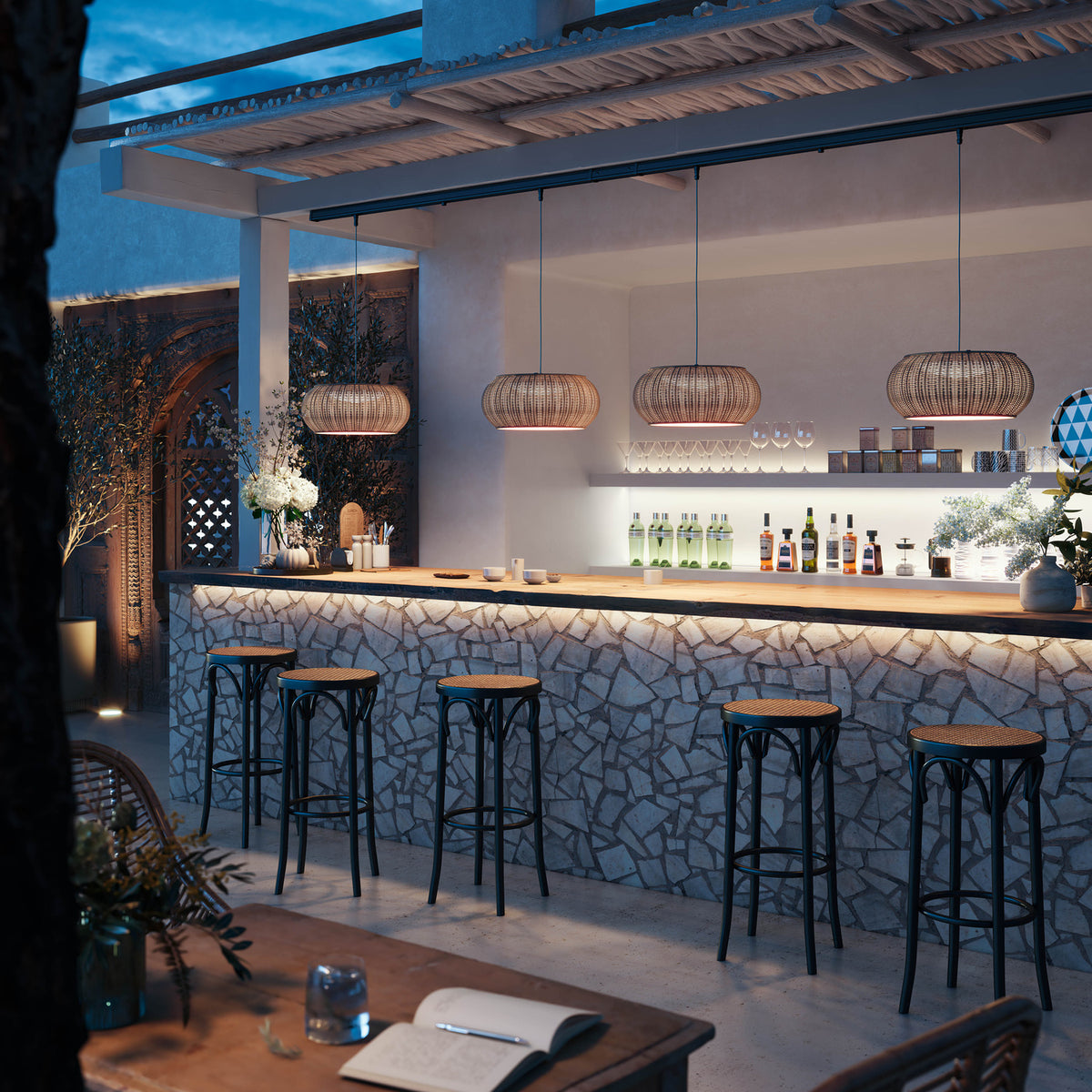 Perris S/47 Outrack woven globe pendant lights in beige hang from an outdoor pergola, illuminating a stone bar with black bar stools at dusk.