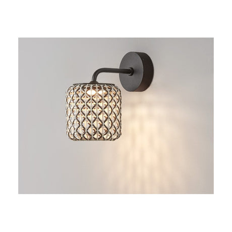 Nans A/01 outdoor wall light with a round woven brown shade and slim bent arm in graphite brown, mounted on a light grey wall and glowing with warm LED light.