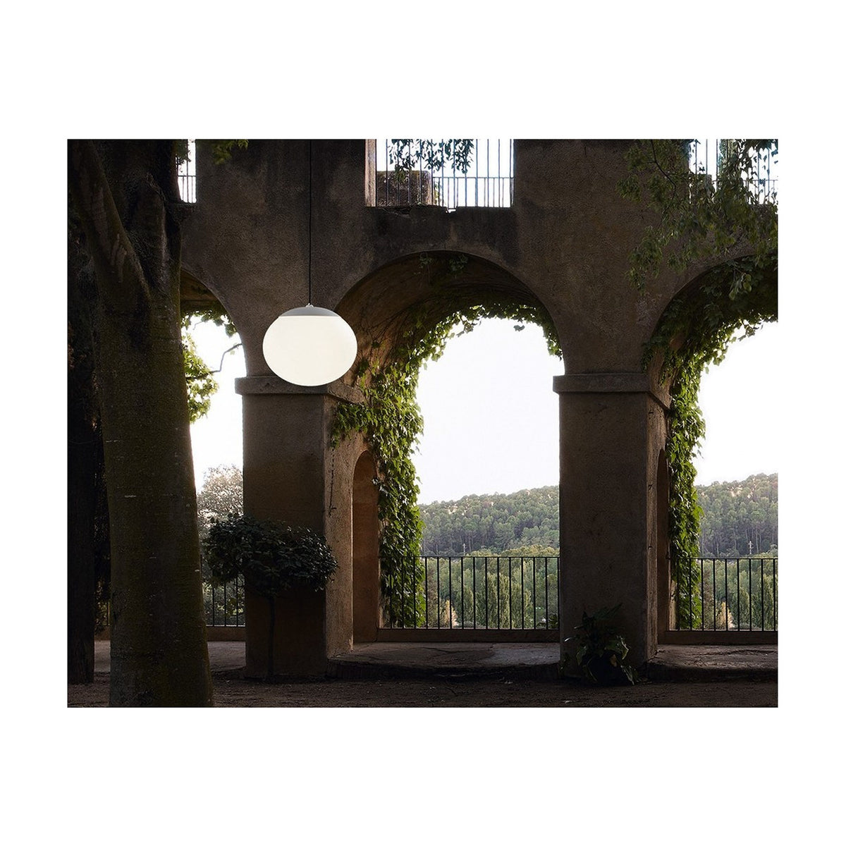 Elipse S/30 white globe LED outdoor pendant light hanging lit beneath a stone archway with climbing greenery and a garden view beyond.