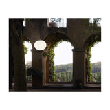 Elipse S/30 white globe LED outdoor pendant light hanging lit beneath a stone archway with climbing greenery and a garden view beyond.