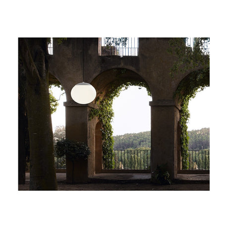 Elipse S/30 white globe LED outdoor pendant light hanging lit beneath a stone archway with climbing greenery and a garden view beyond.