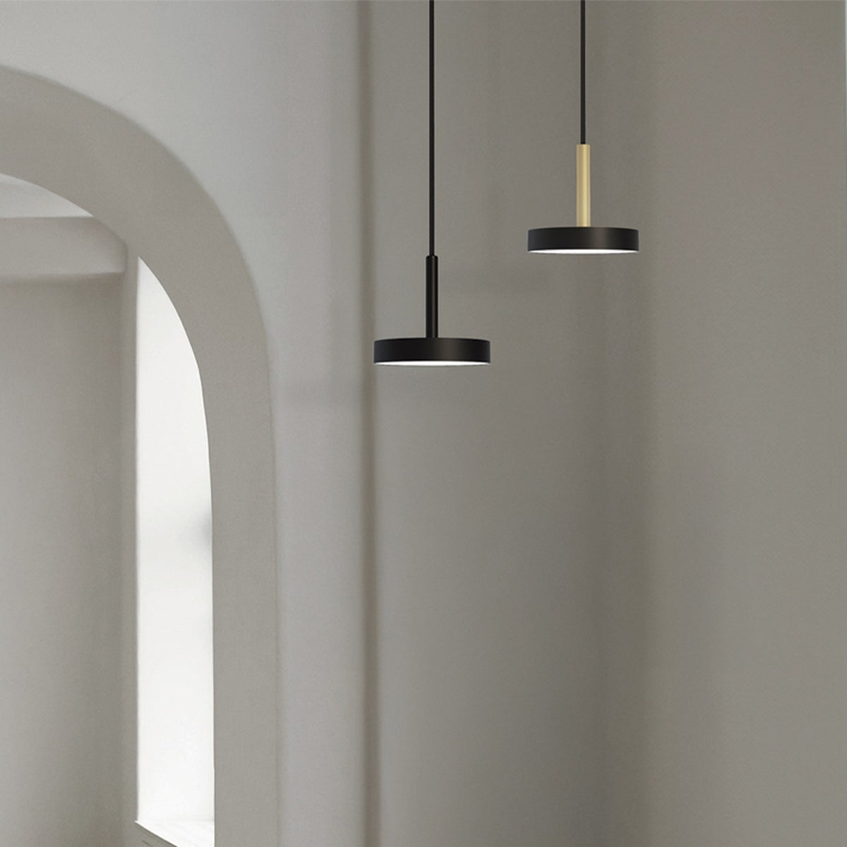 Rafaela Mono LED Drum DALI Pendant in matt black with slim stem and shallow drum shade, shown beside a similar brass-stem pendant in a hallway with arched doorway.