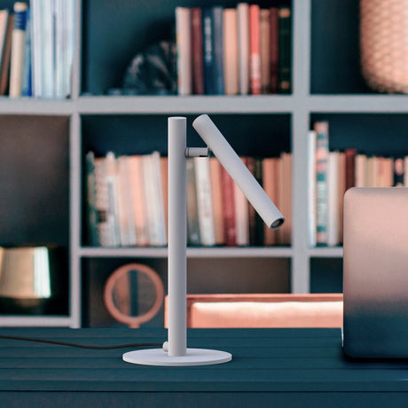 Rui LED Adjustable Reading Table Lamp in matt white, slim upright stem on a round base with an angled tubular shade, shown on a desk against a bookshelf.