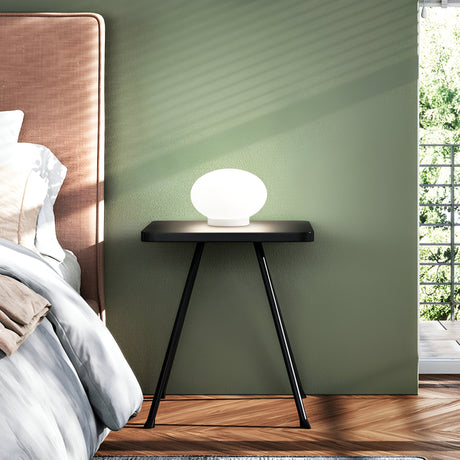 A modern bedroom scene showcases an Orlix Small Table Lamp in white on a sleek black side table next to the bed. The green wall in the room enhances the sunlight streaming through the window, creating shadows. The bed is adorned with white and beige bedding, resting on a wooden floor.