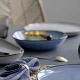 Sandrine Stoneware Deep Plate in blue speckled glaze on a dining table, with grey plates, a linen napkin, brass napkin ring, and spoon in the setting.