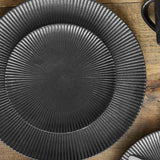 Neri Stoneware Plate in black, round side plate with ridged texture radiating from centre to rim, shown on a wooden surface.