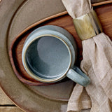 Pixie Stoneware Cup 350ml in green with a wide, low body and open loop handle, shown on a wooden tray beside a folded napkin and horn napkin ring.