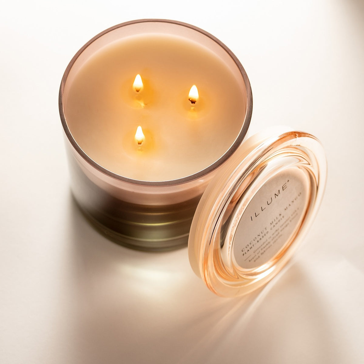 Coconut Milk Mango scented candle in a pink glass vessel with three lit cotton wicks, shown with its glass lid resting beside it on a plain light background.