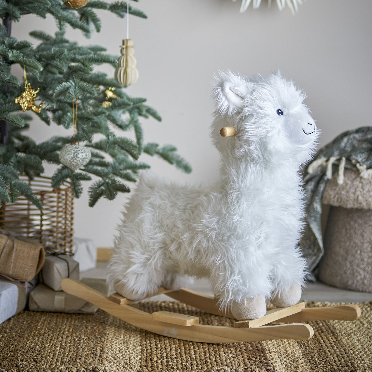 Kinto Rocking Llama in White, a shaggy fabric ride-on toy with wooden handle and curved rockers, shown beside a Christmas tree with ornaments and gifts.
