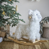 Kinto Rocking Llama in White, a shaggy fabric ride-on toy with wooden handle and curved rockers, shown beside a Christmas tree with ornaments and gifts.