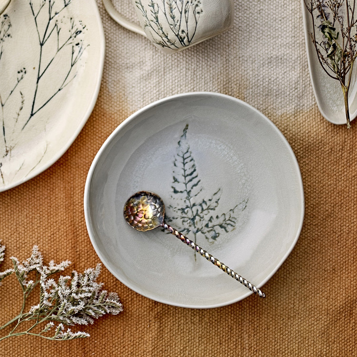 Bea Crackle Stoneware Deep Plate in cream with a fern leaf motif at the centre, styled with a patterned spoon alongside botanical ceramics on a textured tablecloth.