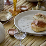 Bea Crackle Stoneware Dinner Plate in cream with floral pattern, shown holding bread rolls beside small bowls, a glass, and a table setting.