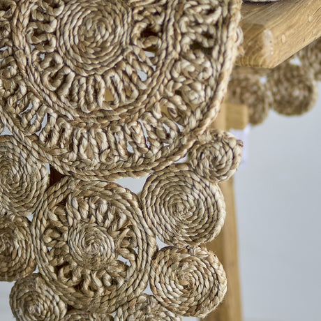 Close-up of the Valder Jute Table Runner in natural jute, showing braided coiled rounds with an openwork spiral pattern, draped over a wooden table edge.