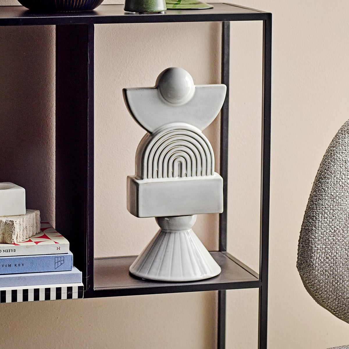 Beatrice Stoneware Ornament in white, a stacked sculptural figure with ball, half-moon, ribbed arch, block and cone shapes, shown on a black shelf unit beside books.