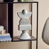 Beatrice Stoneware Ornament in white, a stacked sculptural figure with ball, half-moon, ribbed arch, block and cone shapes, shown on a black shelf unit beside books.