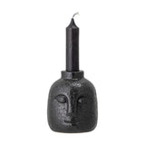 Eliot Stoneware Candle Holder in black, shaped as a face with raised eyes, nose and smile, glossy pitted ceramic finish, holding a slim black taper candle on a white background.