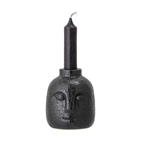 Eliot Stoneware Candle Holder in black, shaped as a face with raised eyes, nose and smile, glossy pitted ceramic finish, holding a slim black taper candle on a white background.