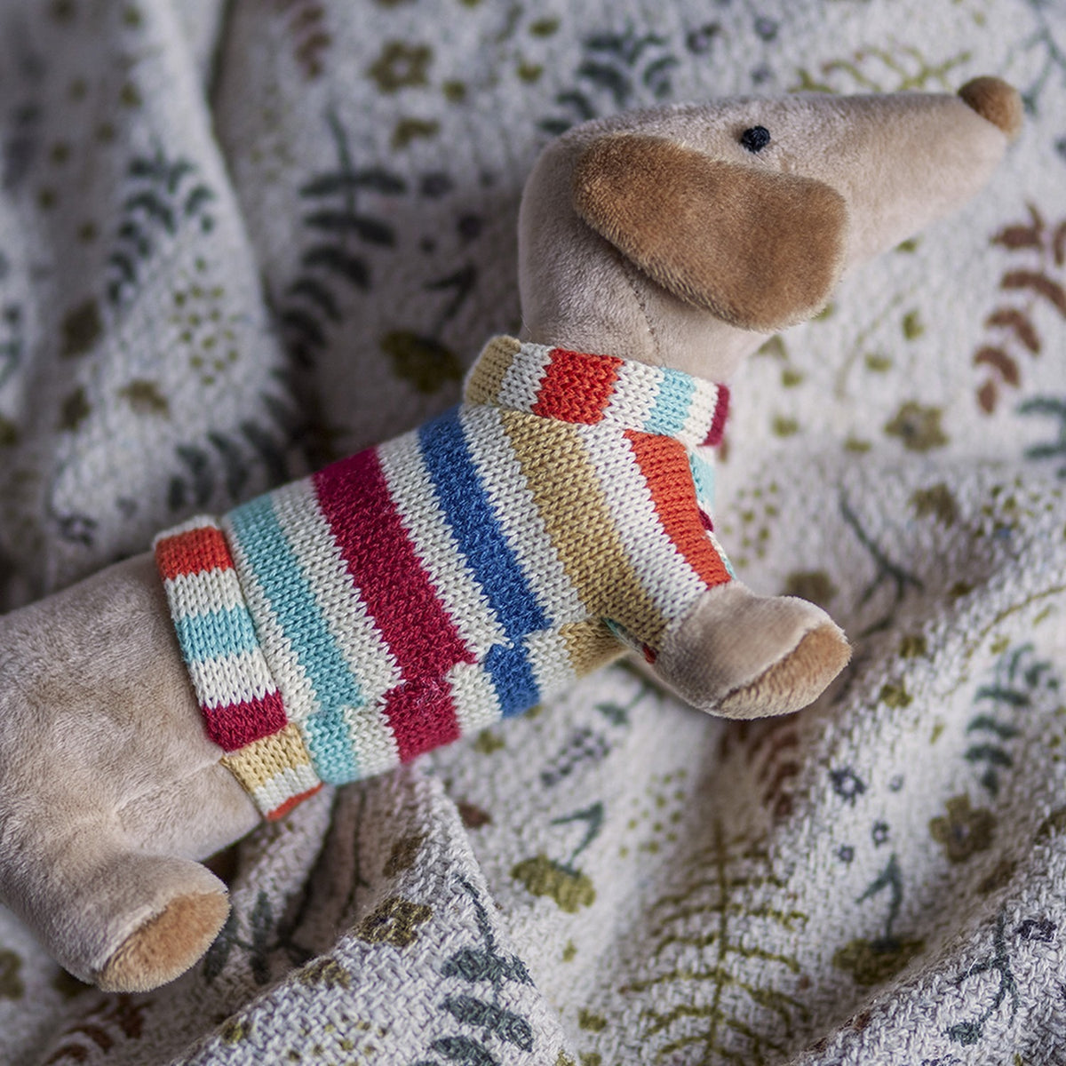 Hilena soft toy sausage dog in a multicoloured striped knitted jumper, shown close-up with floppy ears and rounded nose on a floral patterned fabric background.