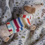 Hilena soft toy sausage dog in a multicoloured striped knitted jumper, shown close-up with floppy ears and rounded nose on a floral patterned fabric background.