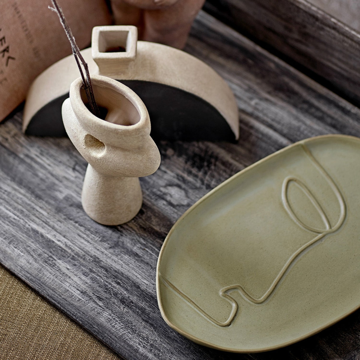 Ngan Stoneware Tray in Green, an oval ceramic tray with raised rim and engraved line-drawn face, shown beside a stoneware vase on a wooden table.